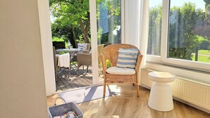 Una silla de mimbre con cojín y almohada, un radiador blanco, y vista a un jardín desde una puerta de cristal.