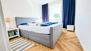Un dormitorio moderno con una cama gris, ropa de cama azul y blanca, una lámpara de noche, una estantería y una ventana con cortinas.