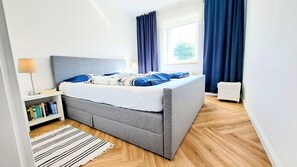A modern bedroom with a grey bed, blue and white bedding, a bedside lamp, a bookshelf, and a window with curtains.