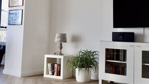 Una sala de estar moderna con un televisor de pantalla plana, una pequeña mesita de noche con una lámpara, una planta en maceta y una estantería.