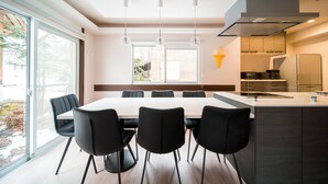A modern kitchen with a central island, a dining area with a table and chairs, and large windows providing natural light. - Yama House Hakuba (Hakuba)