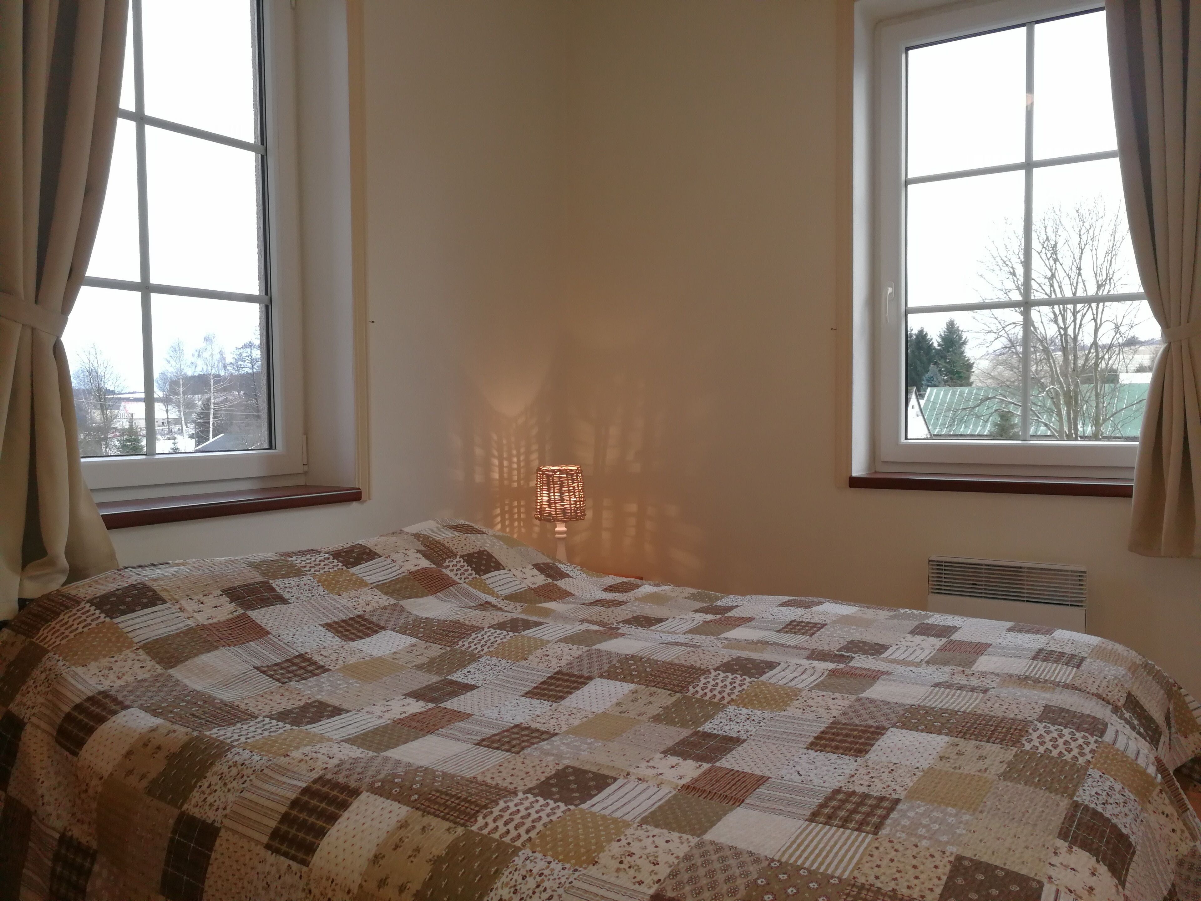 A bedroom with a quilted bedspread, two windows with curtains, and a wall-mounted lamp.