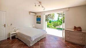Une chambre à coucher comprenant un lit, une table de chevet et une commode. Elle possède un balcon d’où l’on peut admirer la verdure et un immeuble.