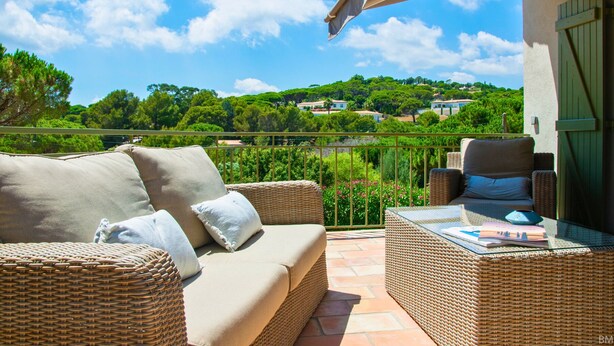 Un balcon agrémenté de meubles en osier, d’une table en verre et offrant une vue sur une colline verdoyante parsemée de maisons.