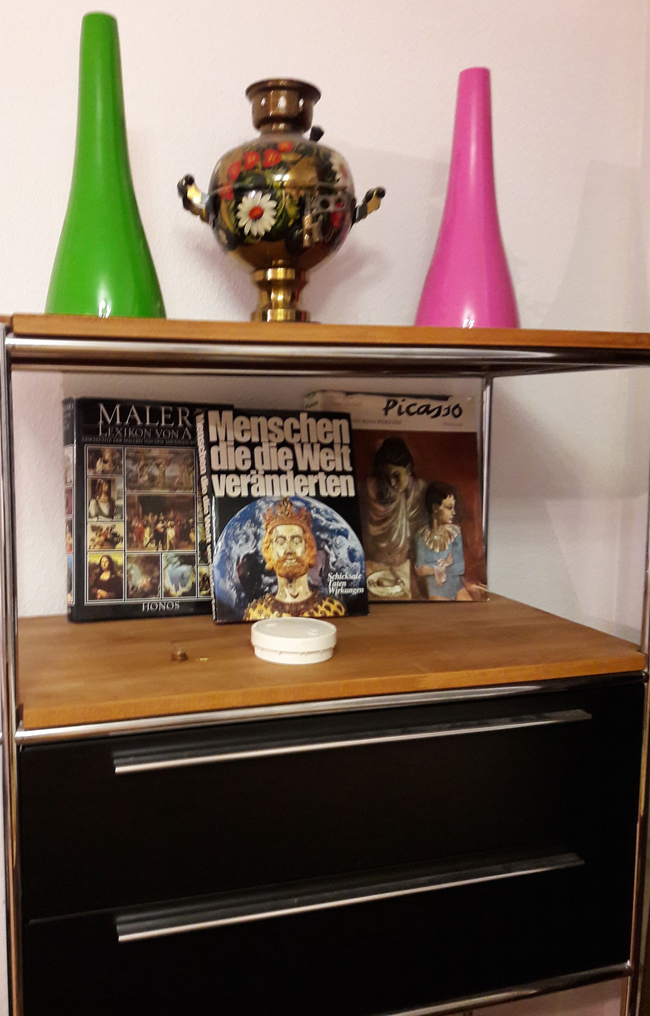 Een plank met decoratieve objecten, waaronder een groene vaas, een roze vaas, een boek getiteld 'Maler Lexikon Vona' en een cd-hoes.