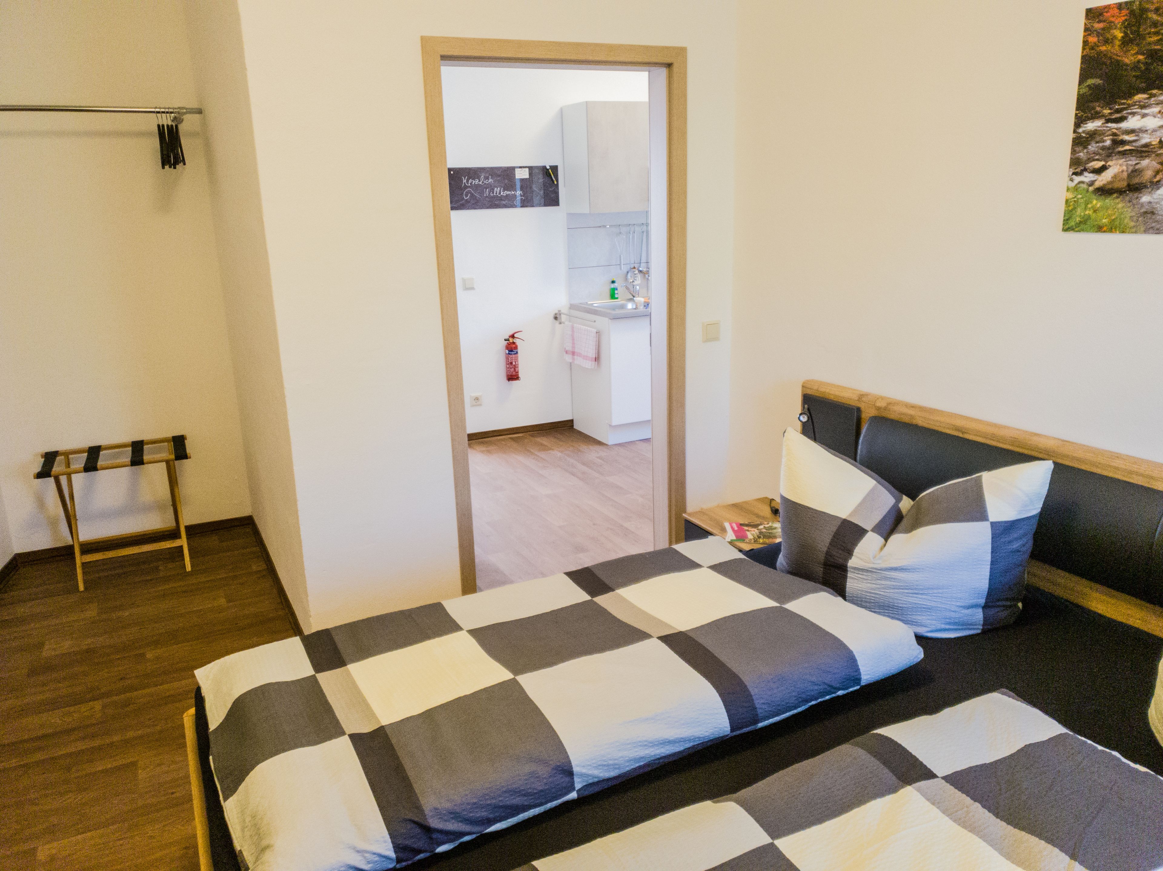 Une chambre à coucher comprenant un lit, une table de chevet, un porte-manteau mural et une porte donnant sur un coin cuisine.