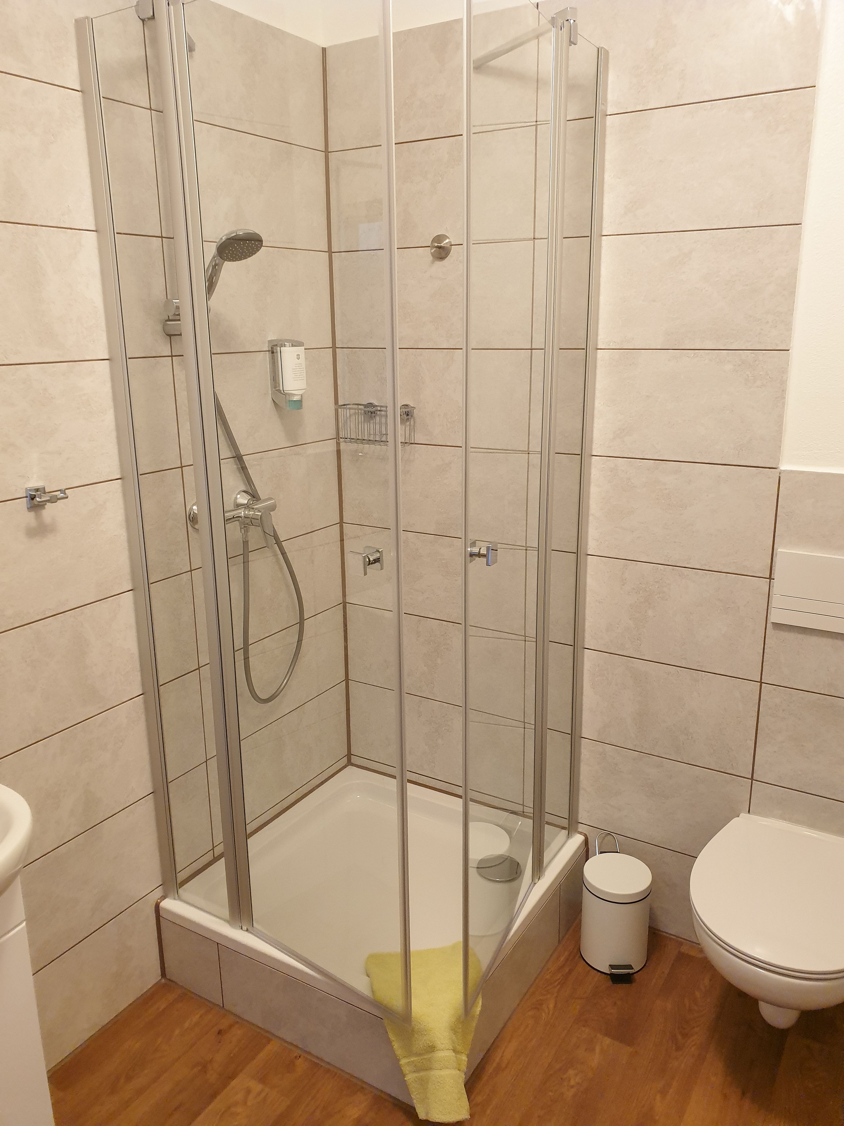 En brusekabine med glasvægge, håndbruser, lille hylde og et hvidt toilet i et badeværelse med fliserede vægge og trægulv.