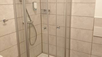 En brusekabine med glasvægge, håndbruser, lille hylde og et hvidt toilet i et badeværelse med fliserede vægge og trægulv.