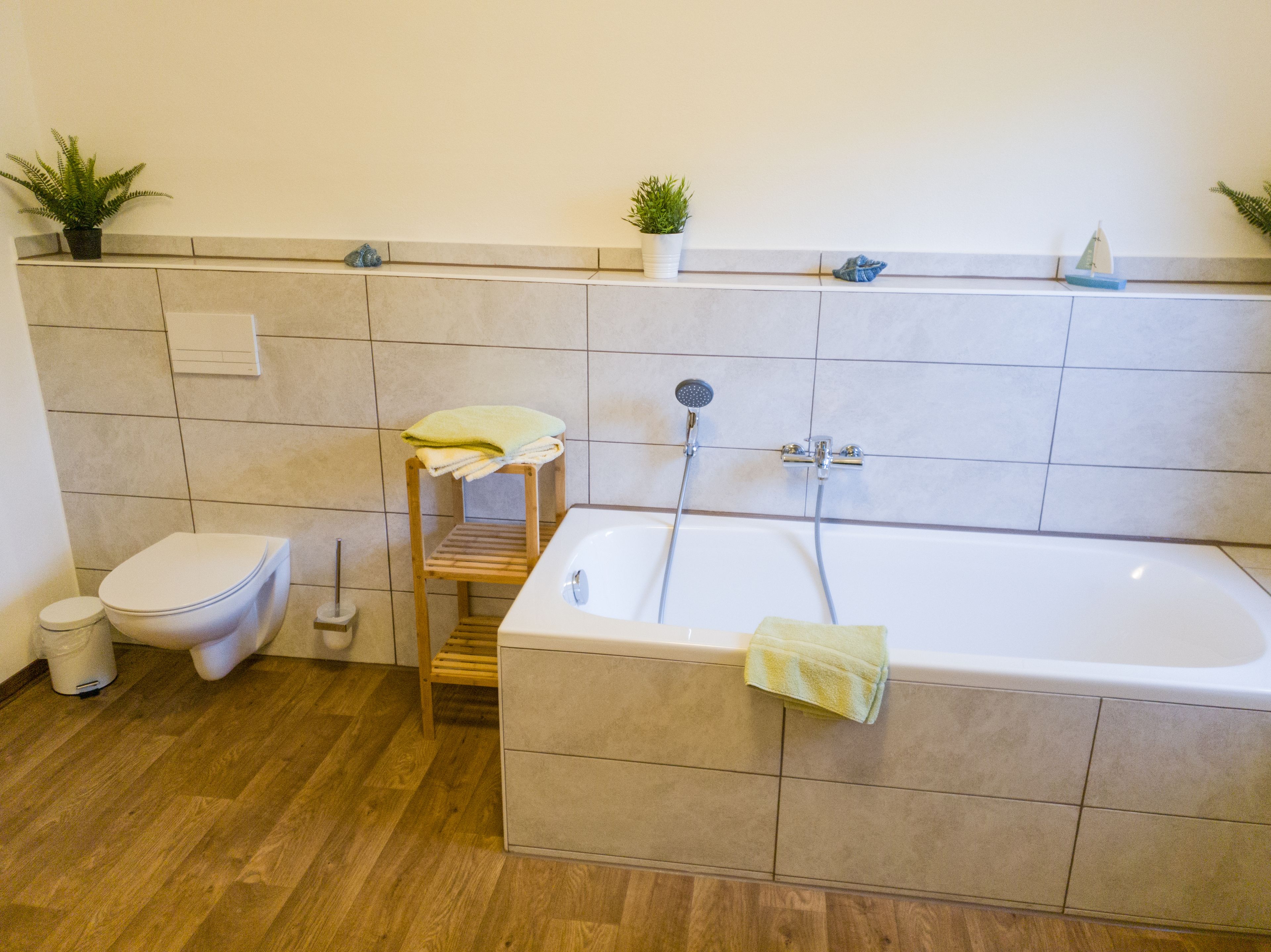 A bathroom with a toilet, a wooden shelf with towels, a bathtub with a showerhead, and two potted plants on the wall.