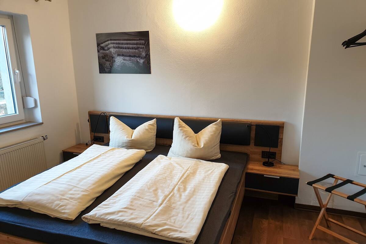 A neatly made bed with two pillows, a bedside table with a lamp, and a wall-mounted picture.