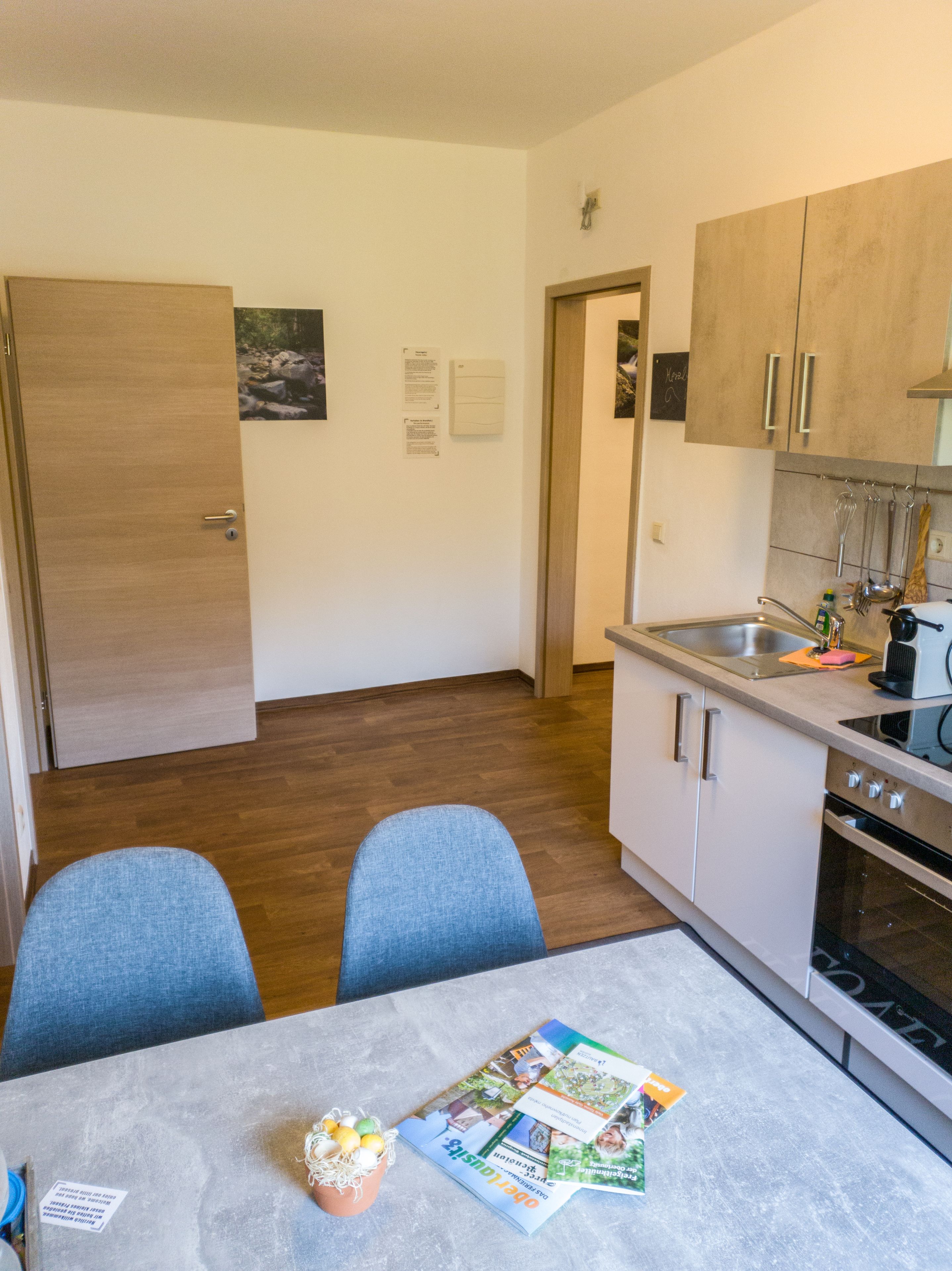 Cocina compacta con armarios de madera, un lavamanos y una estufa. Hay una mesa de comedor con dos sillas y una mesita con una revista y un objeto decorativo.