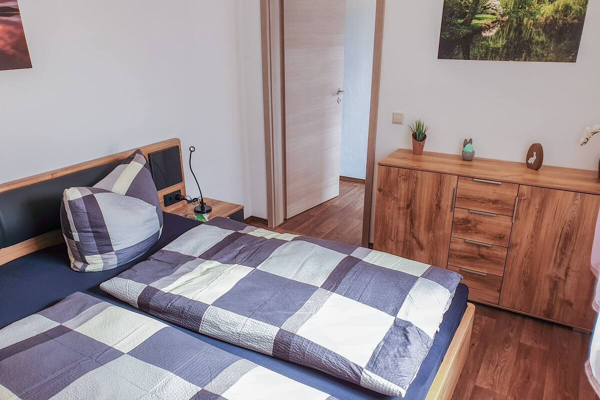 A bedroom with a bed, wooden nightstand, dresser, and a door leading to another room.