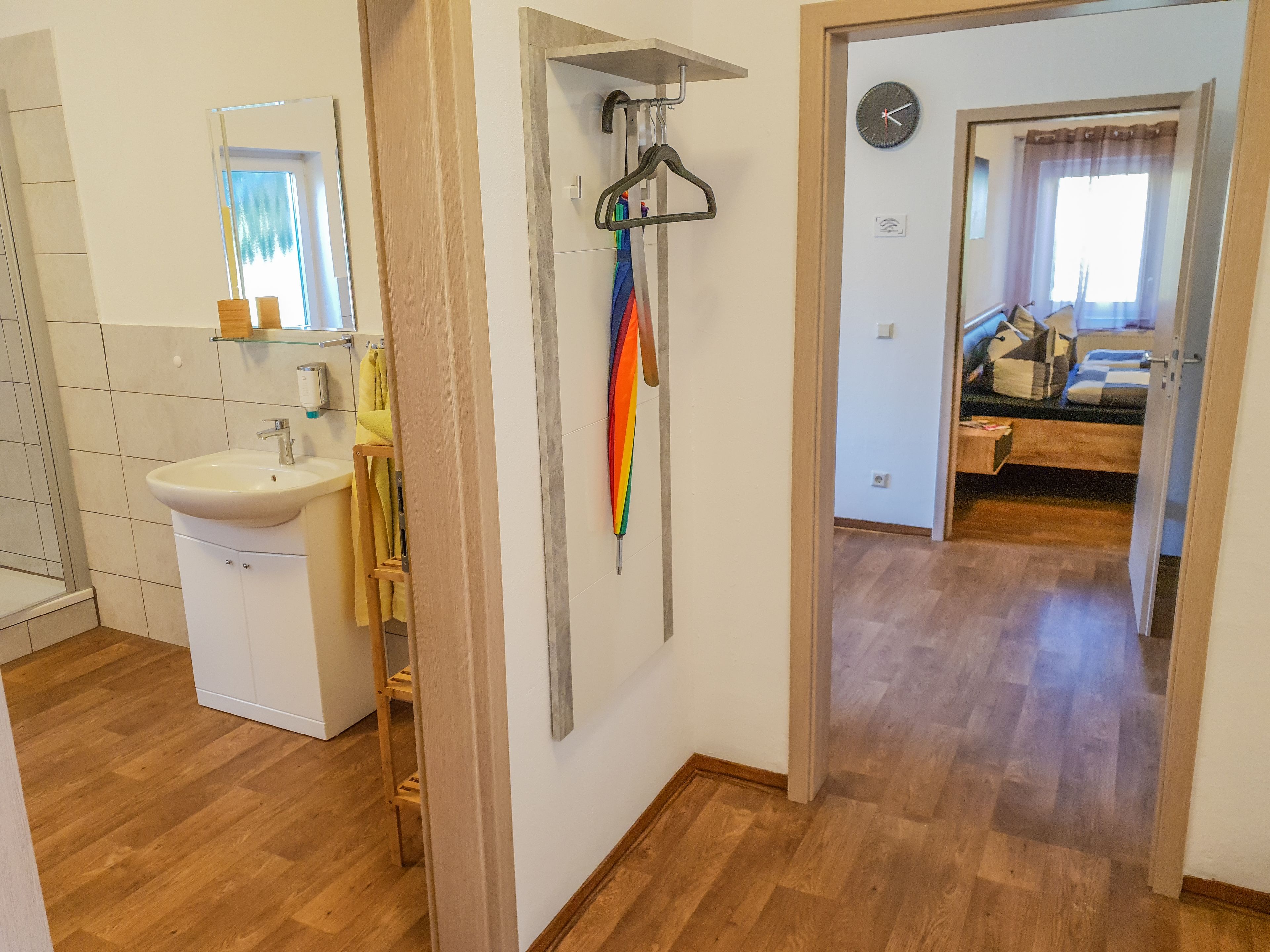 A bathroom with a sink, mirror, and towel rack. A hallway leads to a bedroom with a bed and a clock.