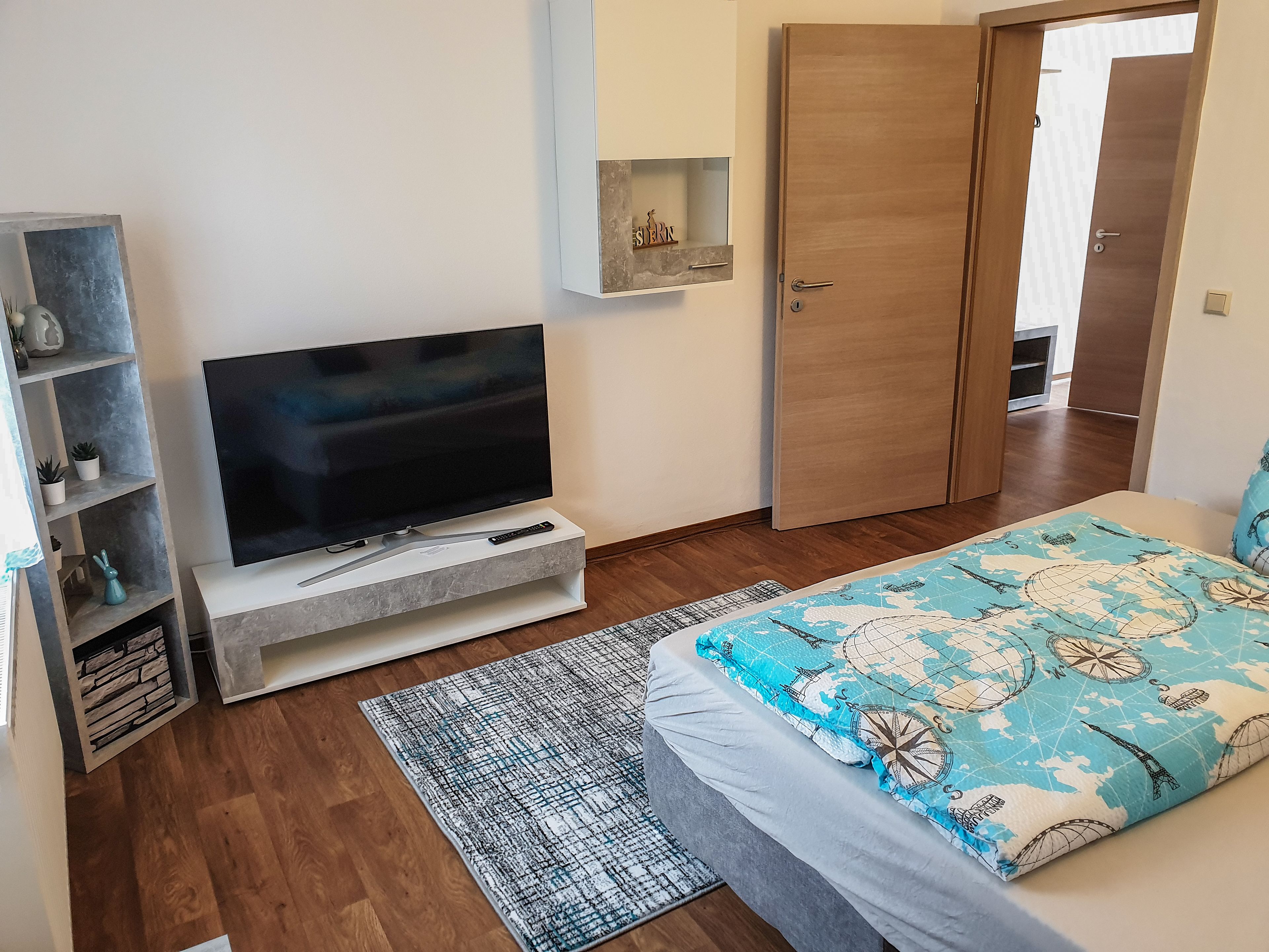 Un dormitorio con una televisión sobre un mueble blanco, una puerta de madera, una cama con colcha azul y blanca estampada y una alfombra estampada sobre el suelo de madera.