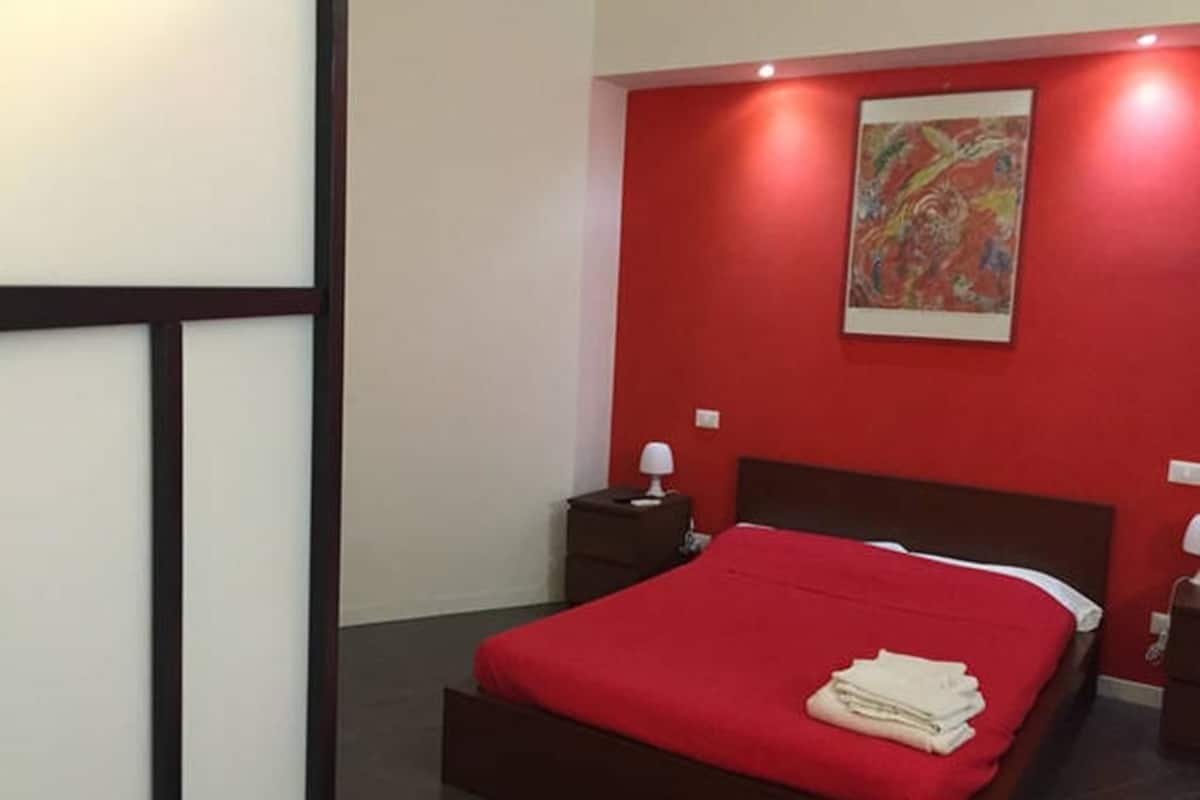 A bedroom with a red wall, a bed with a red bedspread, a nightstand with a lamp, and a painting above the bed.