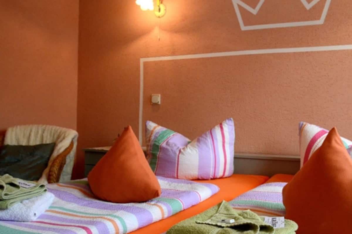 A hotel room with two beds, each with striped bedding and orange pillows. A crown-shaped light fixture is on the wall above the beds.