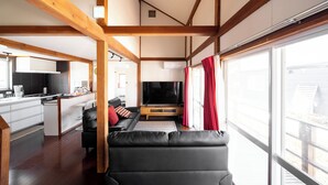 A modern living room with a black sofa, wooden beams, a kitchen area, and a large window with curtains. - Hanna's House Hakuba (Hakuba)