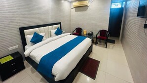 A modern hotel room with a large bed, a ceiling fan, an air conditioning unit, a TV, and a small seating area with chairs. - Hotel Gangotri (Haridwar)