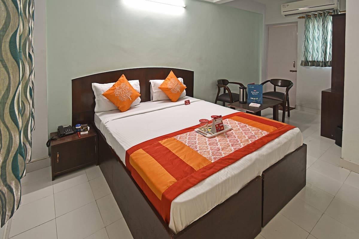 A neatly made bed with a patterned bedspread, a bedside table with a phone and a book, and a chair in the corner.