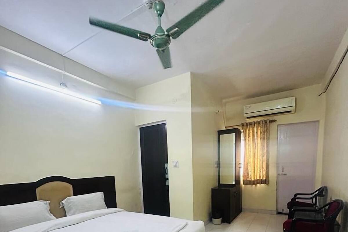 A hotel room with a bed, a ceiling fan, an air conditioning unit, a mirror, a chair, and a small table.