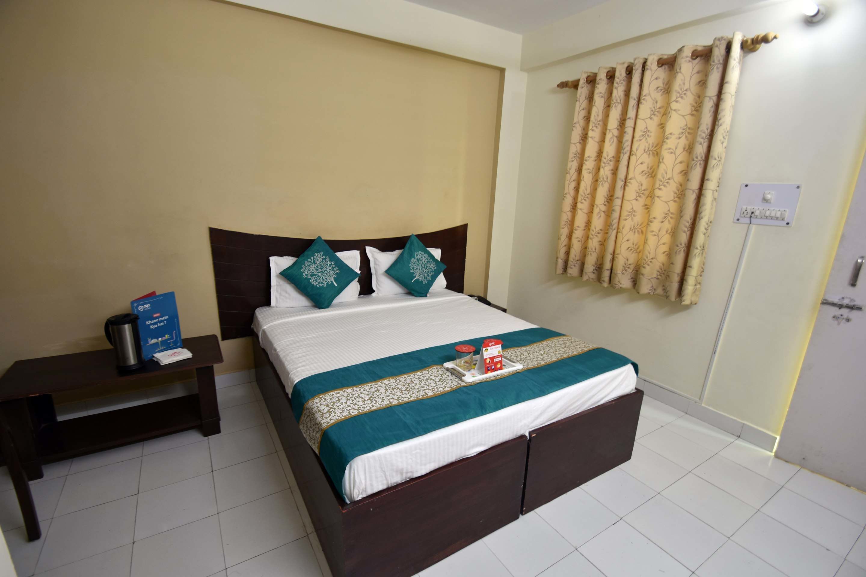 A neatly made bed with a white and teal bedspread, two teal pillows, and a bedside table with a coffee maker and a box.