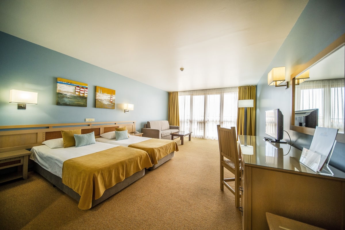 A hotel room with two beds, a desk, a chair, a TV, and artwork on the walls.