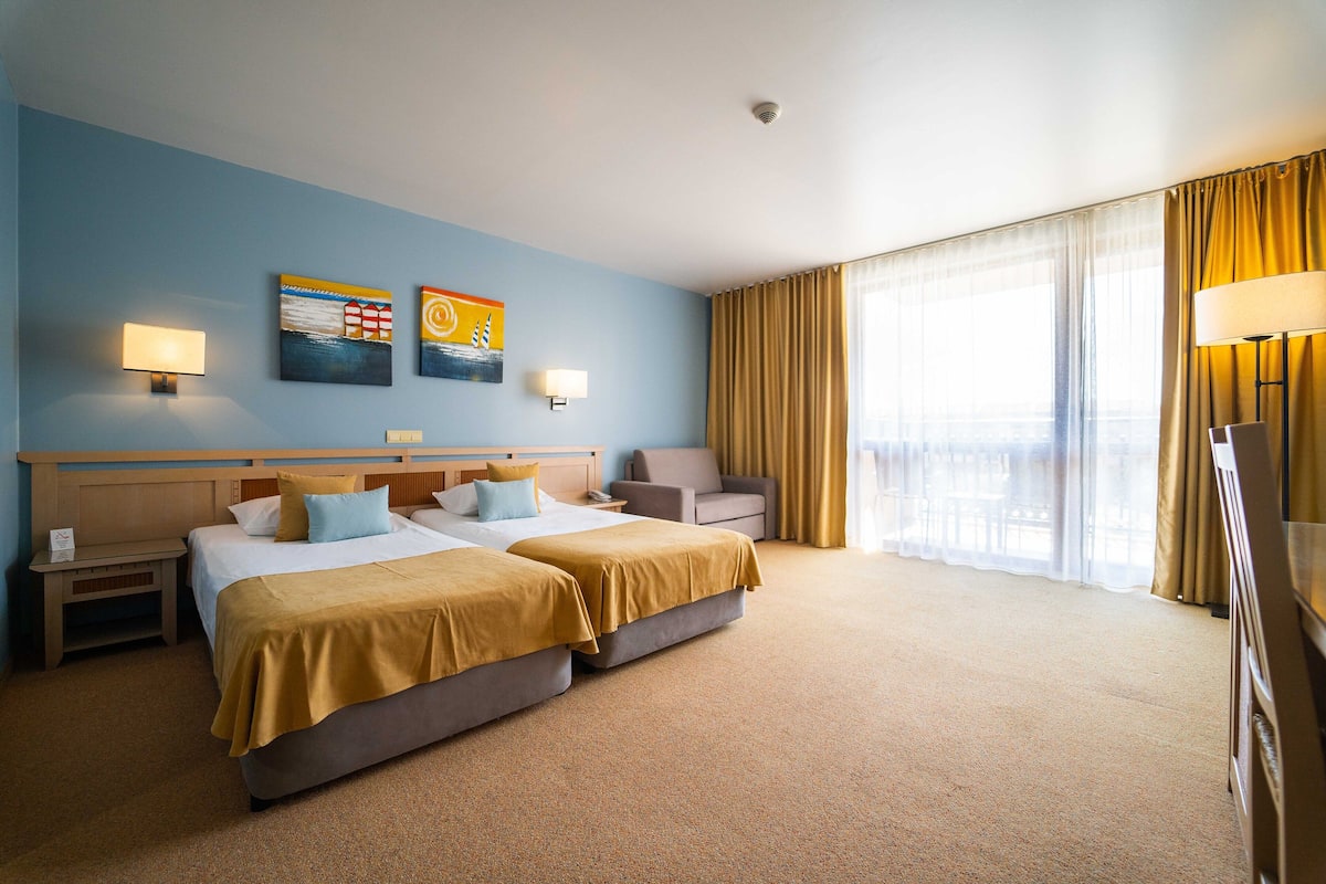 A hotel room with two beds, a desk, a chair, a TV, and a large window with curtains.