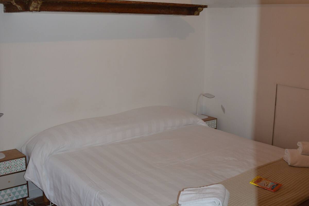 A single bed with white linens, a bedside table with a lamp, and a small bedside table with a book.