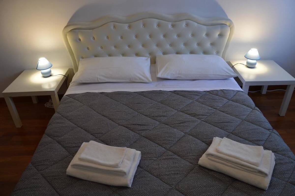 A neatly made bed with a grey quilt, two white pillows, and folded towels on the bed. There are two bedside tables with lamps on either side of the bed.