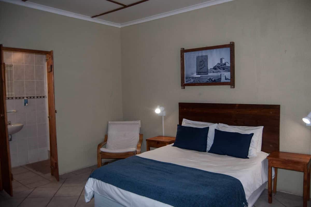 Une chambre d’hôtel avec un lit, une chaise, une table de chevet, un tableau accroché au mur et une porte de salle de bain.
