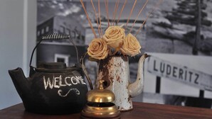 A black teapot with "WELCOME" written on it, a white vase with roses, and a brass bell. - Obelix Guesthouse (Luderitz)
