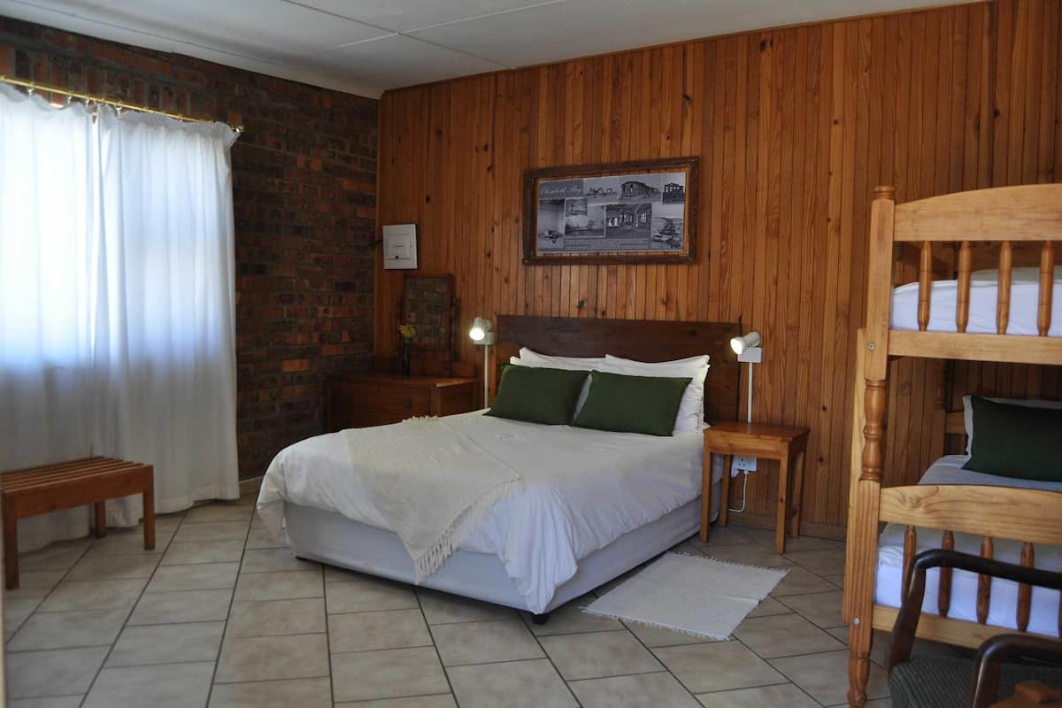 Une chambre avec une tête de lit en bois, un lit simple avec une literie blanche, une table de chevet avec une lampe, un banc et un mur en briques.