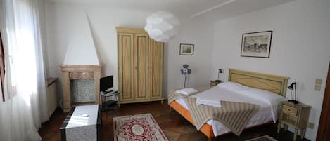 Un dormitorio con cama, mesitas de noche, escritorio, silla, armario, chimenea y una lámpara en el techo.