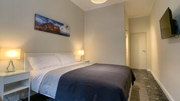 A hotel room with a bed, two bedside lamps, a painting on the wall, a television mounted on the wall, and a door leading to another room.