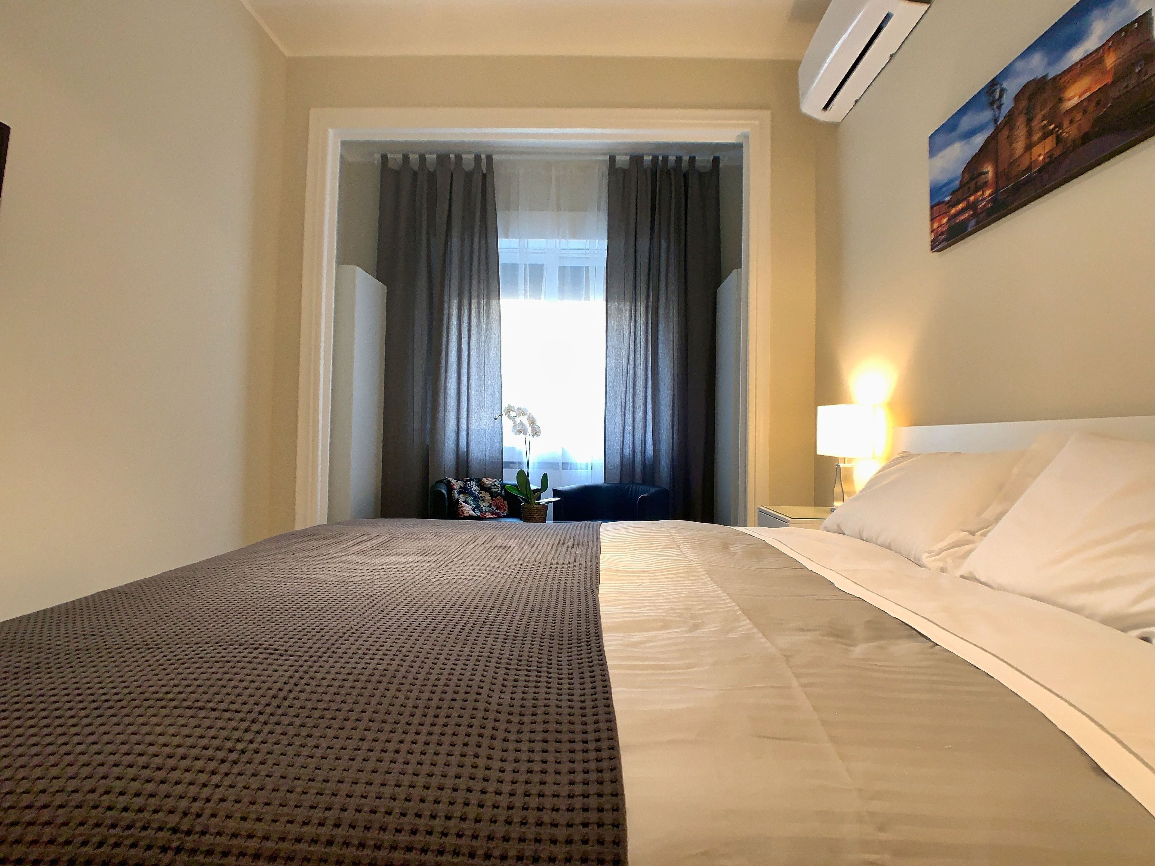 Una habitación de hotel con una cama grande, una mesita de noche, una lámpara, una ventana con cortinas y un cuadro en la pared.