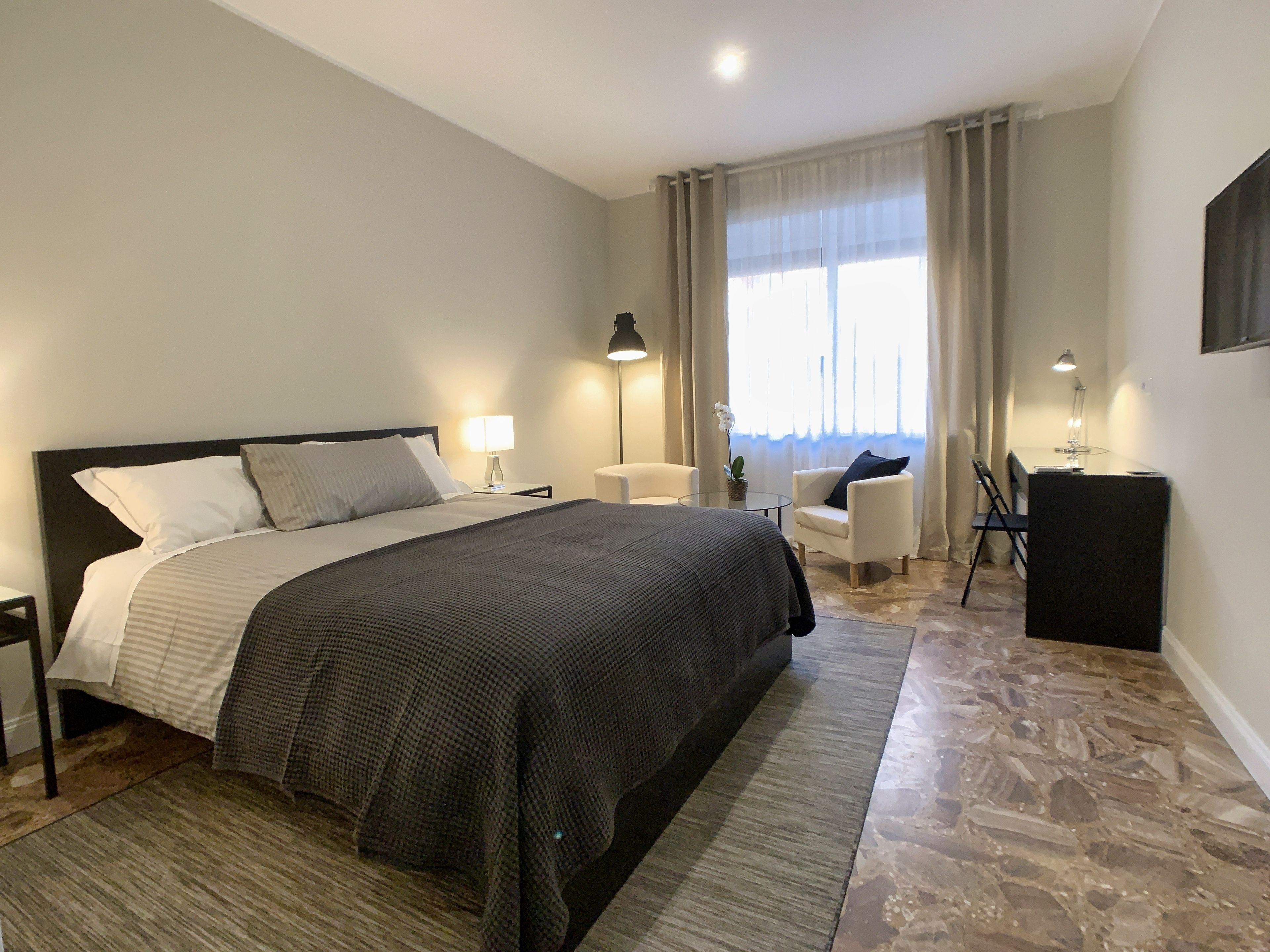 A modern hotel room with a large bed, a desk, a chair, and a small table.