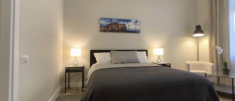 A neatly arranged bedroom with a large bed, two bedside tables with lamps, a chair, and a small table with a plant.