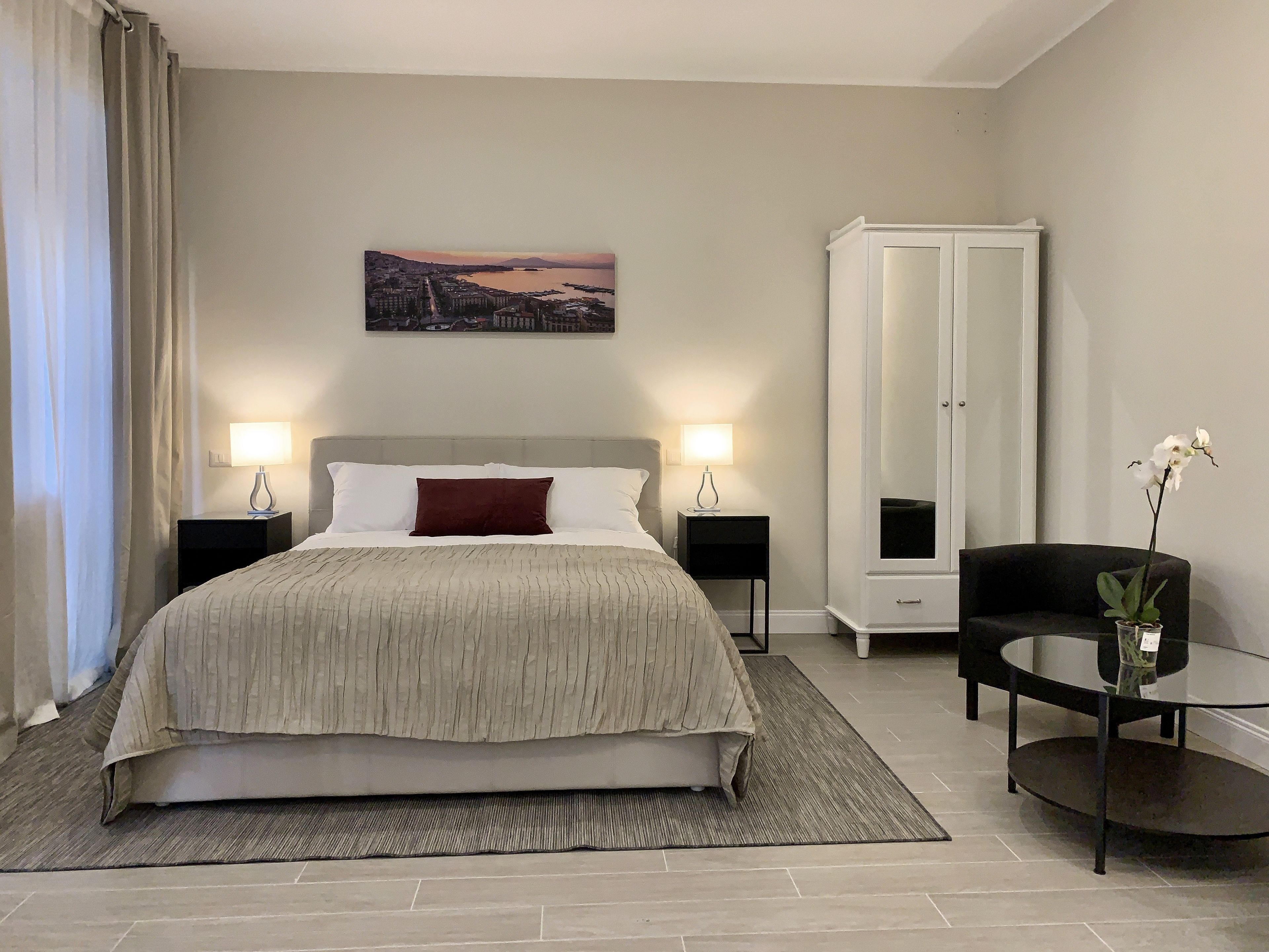A hotel room with a large bed, two bedside tables with lamps, a wardrobe, a small table with a vase, and a painting on the wall.