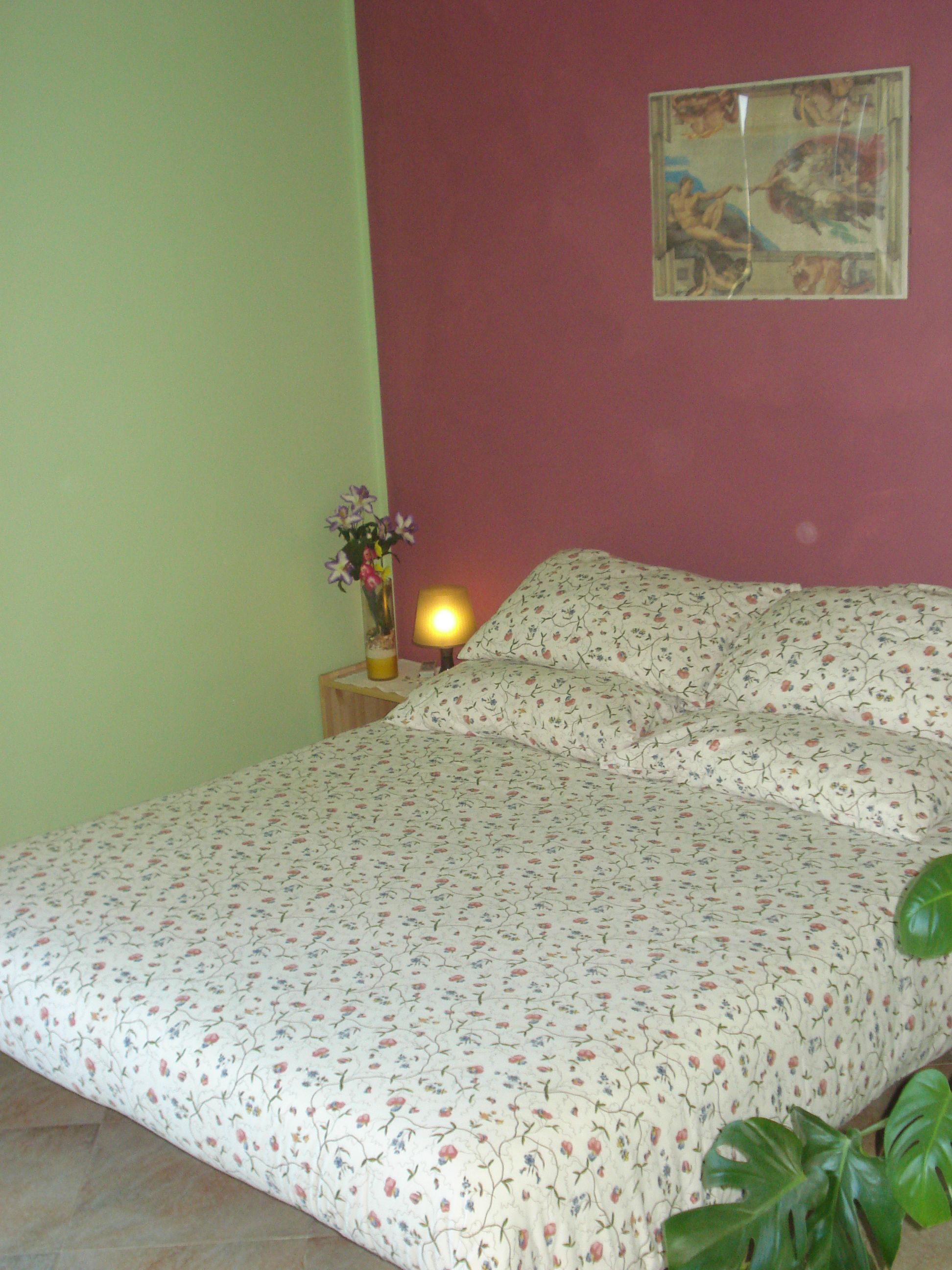 Een slaapkamer met een bloemenbedekker, een nachtkastje met een lamp, een vaas met bloemen en een schilderij aan de muur.