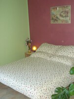 A bedroom with a floral bedspread, a nightstand with a lamp, a vase with flowers, and a painting on the wall.