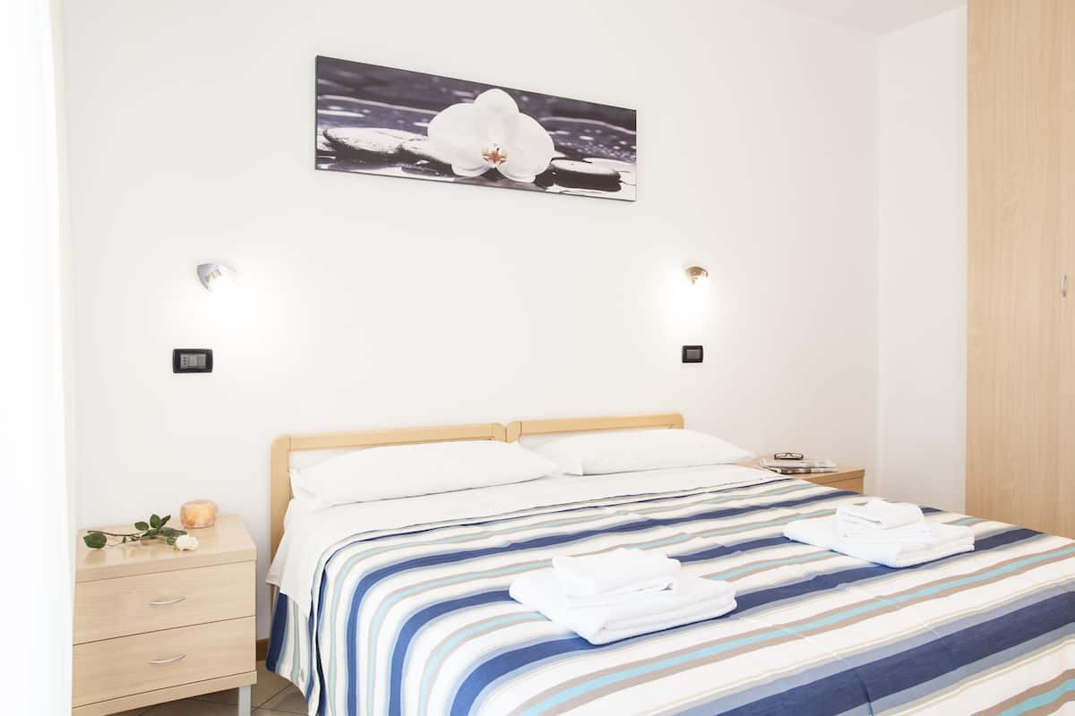 Una cama bien hecha con ropa de cama a rayas, una mesita de noche con lámpara y un cuadro de una orquídea colgado en la pared.