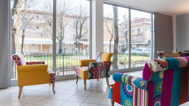 A spacious room with large windows, a yellow armchair, a colorful sofa, and a small table. - Residence Cigno (Rimini)