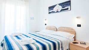 A hotel room with a bed, a nightstand, a wall-mounted painting, and a window with sheer curtains. - Residence Cigno (Rimini)