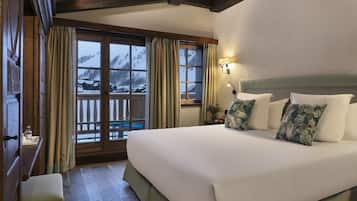 Une chambre d’hôtel avec un grand lit, des poutres apparentes, un balcon offrant une vue sur des montagnes enneigées et une table de chevet avec une lampe.