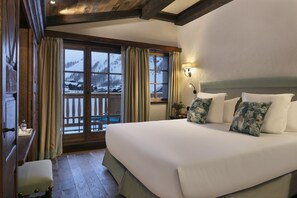 A hotel room with a large bed, wooden beams, a balcony with a view of snow-covered mountains, and a nightstand with a lamp.