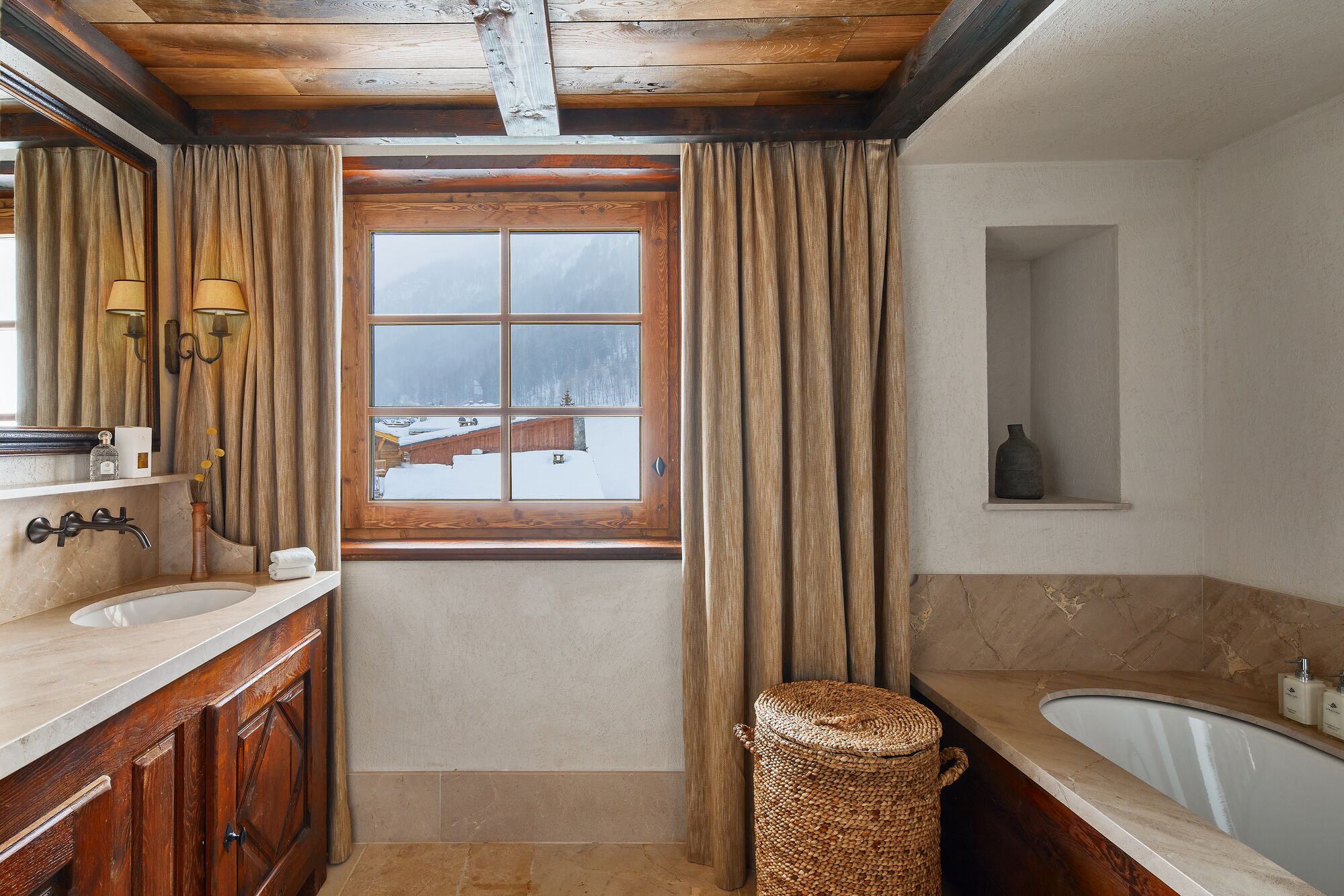 A bathroom with a wooden ceiling, a window with curtains, a sink, a bathtub, and a woven stool.