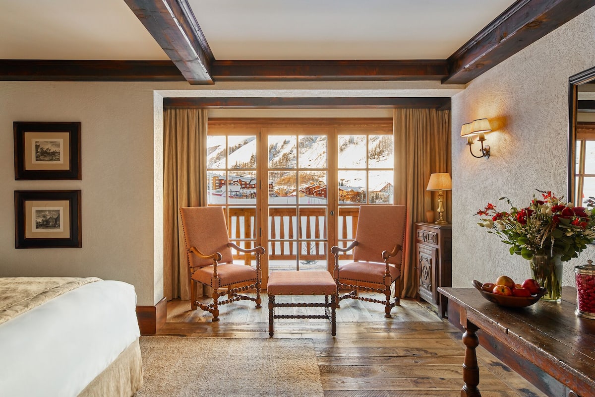 A room with a large window, two chairs, a wooden bench, a bedside table with a vase of flowers, and a framed picture on the wall.