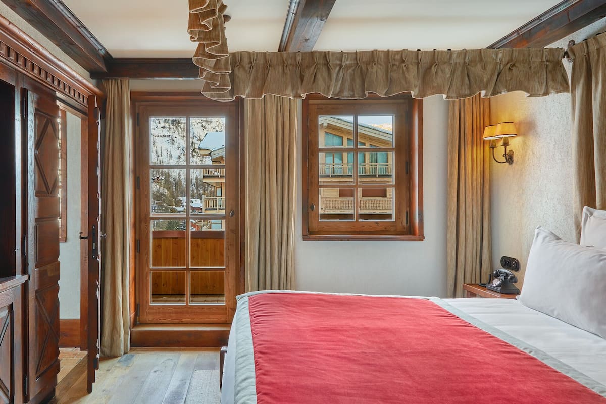A room with a large bed, a wooden door with glass panels, a window with a view of a building, and a wall-mounted lamp.