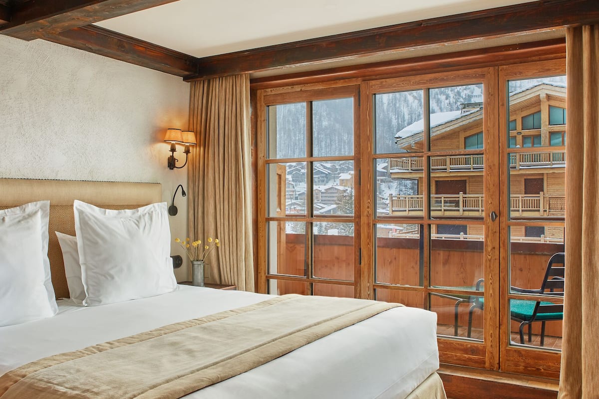 A hotel room with a large bed, wooden walls, a balcony with a view of a snowy mountain, and a wooden railing.