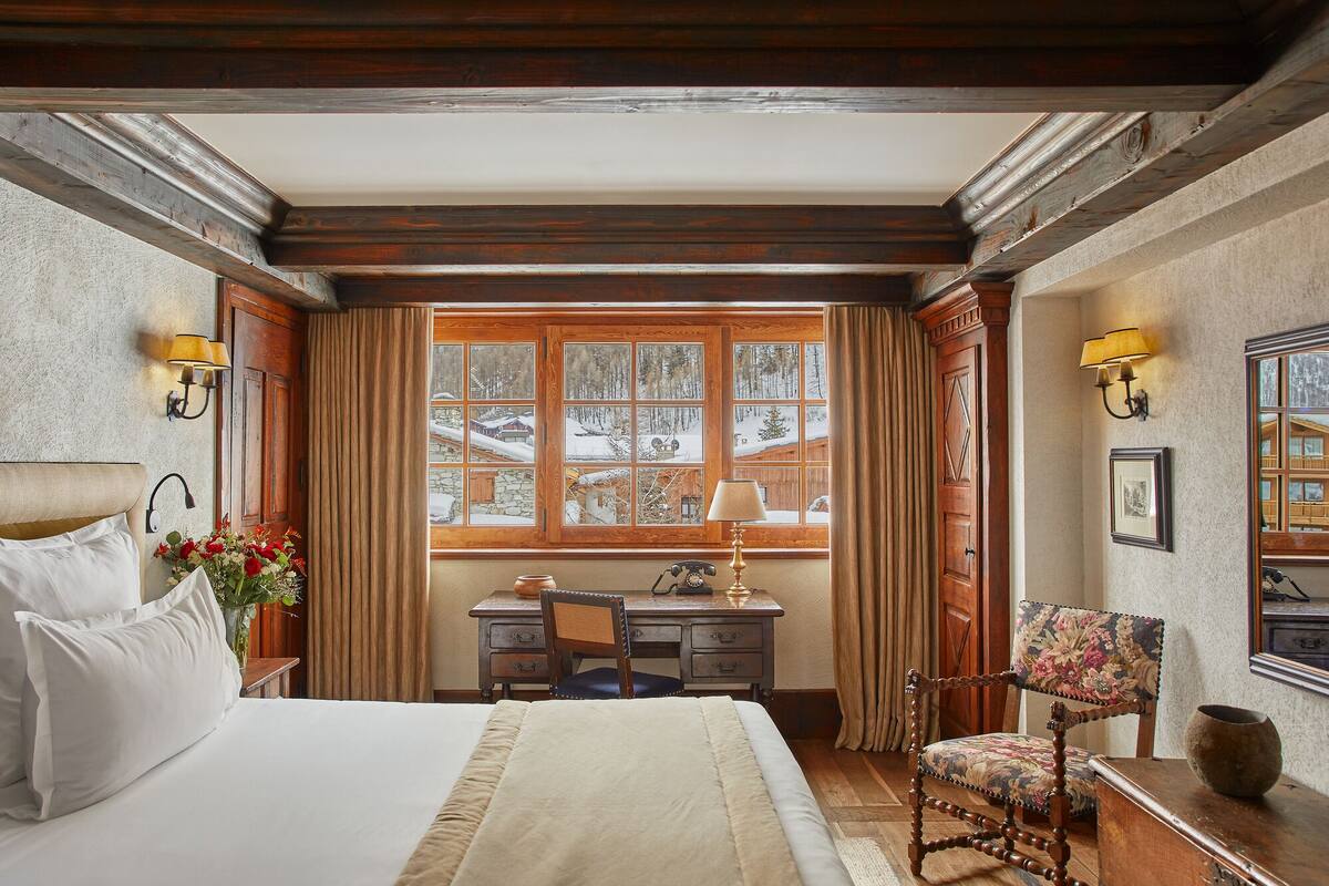 A hotel room with a large bed, a desk with a telephone, a chair, and a window with a view of a snowy landscape.